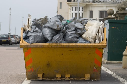 A large yellow industrial skip filled with numerous black and grey plastic rubbish bags, some appearing slightly crumpled and others more tightly sealed. The skip is positioned on a paved surface, with orange and red reflective safety markings on its sides. Behind the skip, structures typical of a seaside town can be seen, including a white building with bay windows and a green waste collection container. The surrounding environment suggests an urban street setting, possibly in a residential or commercial area within the vicinity of Soho House, with parked cars visible in the background and a cloudy sky overhead. The scene depicts a rubbish removal service, with clear emphasis on the filled skip ready for collection, consistent with professional waste disposal standards in the London area, such as Soho House postcode.