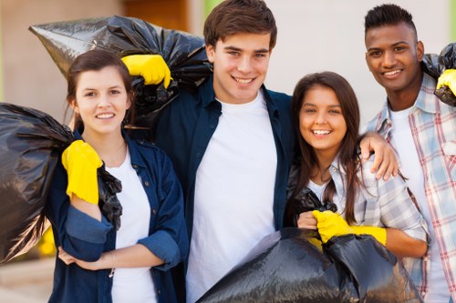 Photograph of four young adults participating in a community rubbish removal activity outdoors, standing close together with bright smiles. They are dressed casually, with some wearing yellow work gloves, and are holding large black rubbish bags filled with waste. The background shows a paved outdoor space, possibly a residential or urban area, with blurred buildings and greenery. The individuals appear engaged and positive, contributing to local cleanliness efforts, which relates to rubbish disposal services. The setting emphasizes teamwork in clearing litter or waste, aligning with professional rubbish removal activities in the area, such as Soho House, near London. The scene is well-lit with natural daylight, highlighting their friendly expressions and the collected rubbish, reinforcing the context of environmental cleanup and waste management support in a community setting.