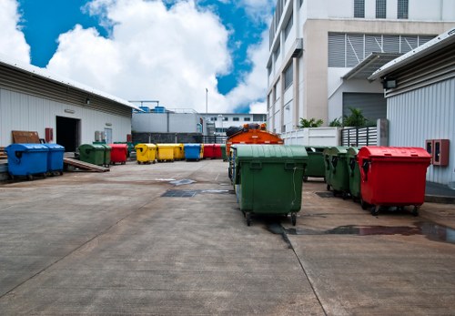 A spacious outdoor area within an industrial or commercial site, featuring multiple large rubbish bins in bright colours including green, red, yellow, and blue, lined up along a concrete ground. The bins have sturdy, smooth plastic textures with some showing metal handles and lids, indicating they are used for waste collection. Behind the bins, there are two white warehouse-style buildings with metal sidings, one on the left and another in the background, with a clear blue sky and scattered white clouds overhead. To the right, part of an adjacent building is visible, with horizontal metal cladding and window blinds. The area appears clean, well-organized, and suitable for rubbish collection and disposal services, consistent with the environment in Soho, London, where Soho House Clearance operates. The scene suggests a professional waste management setting, suitable for customer site clearance or rubbish removal, with a focus on practical, high-capacity waste receptacles.